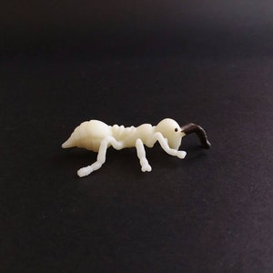 May include: A small, off-white ant figurine with a dark brown head and antennae. The ant is posed on a black surface, showcasing its detailed body structure and delicate legs. This miniature insect model is a decorative item.