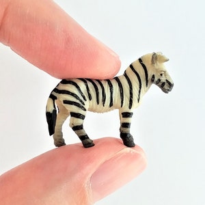 Può includere: Una piccola statuina di zebra di plastica con strisce bianche e nere. La zebra è in piedi su un dito.