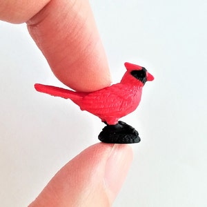 Puede incluir: Una pequeña figura de cardenal rojo con ojos negros y pico negro. La figura está de pie sobre una base negra.