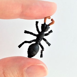 May include: A small, black ant figurine with brown antennae. The ant has six legs and a segmented body. The figurine is held between two fingers against a white background.