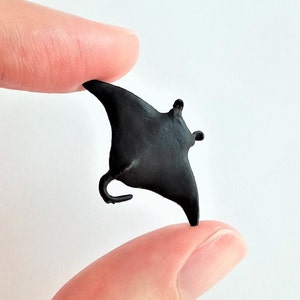 May include: A small, black, plastic manta ray figurine. The figurine is about the size of a finger and is held between two fingers.