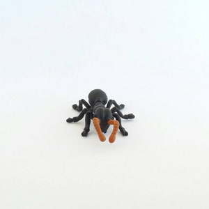 May include: A black plastic ant figurine with orange antennae and mandibles. The ant has six legs and is positioned on a white surface. This insect model is a detailed representation of an ant.