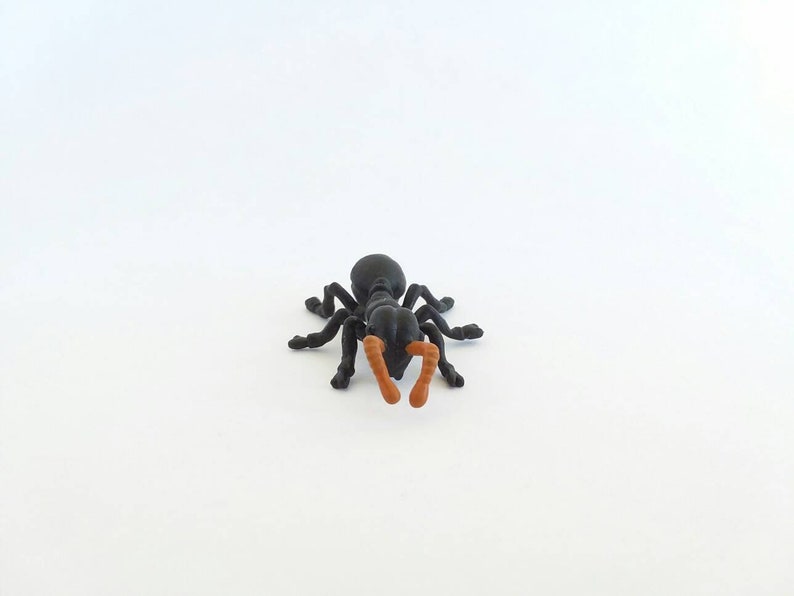 May include: A black plastic ant figurine with orange antennae and mandibles. The ant has six legs and is positioned on a white surface. This insect model is a detailed representation of an ant.