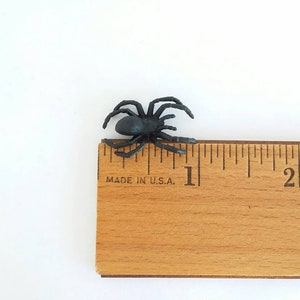 May include: A black plastic spider measuring approximately 1 inch in length. The spider is placed next to a wooden ruler with the text "MADE IN U.S.A."