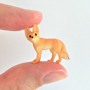 May include: A miniature orange fox figurine with large ears and black eyes. The small fox is standing on a finger, showcasing its detailed features and small size. The figurine has a light orange body and a black-tipped tail.