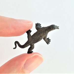 May include: A small, black plastic toy lizard with a textured surface. The lizard is posed in a standing position with its tail curled.
