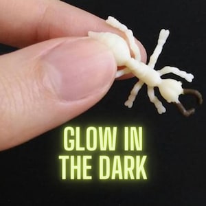 May include: A small, off-white ant figurine with brown pincers is held between fingers against a black background. The text "GLOW IN THE DARK" is displayed in bright green, indicating a special feature of the toy.