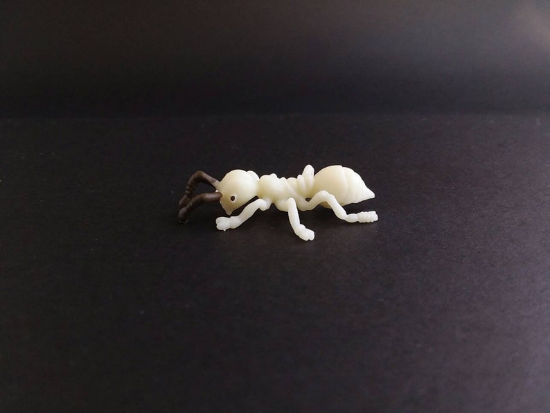 May include: A small, off-white plastic ant figurine with dark brown antennae and eyes. The ant is posed on a black surface, showcasing its segmented body and legs. This decorative item could be used for educational purposes or as a novelty.