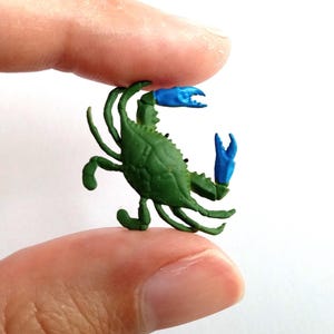 May include: A small, green crab figurine with blue claws. The crab is held between two fingers, showcasing its detailed design and vibrant colors. The figurine is a miniature representation of a crab.