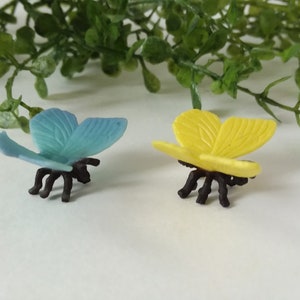 Peut inclure: Deux figurines de papillons en plastique, l'une bleue et l'autre jaune, avec des pattes et des antennes noires. Les papillons sont pos&eacute;s avec leurs ailes ouvertes.