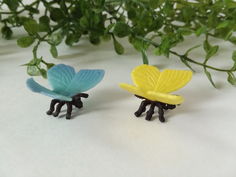 Peut inclure: Deux figurines de papillons en plastique, l'une bleue et l'autre jaune, avec des pattes et des antennes noires. Les papillons sont pos&eacute;s avec leurs ailes ouvertes.