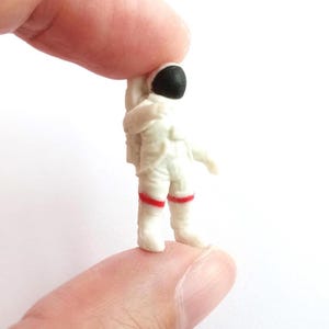 May include: A miniature astronaut figurine in a white spacesuit with a black helmet and red accents. The small figure is held between fingers, showcasing the details of the space suit.