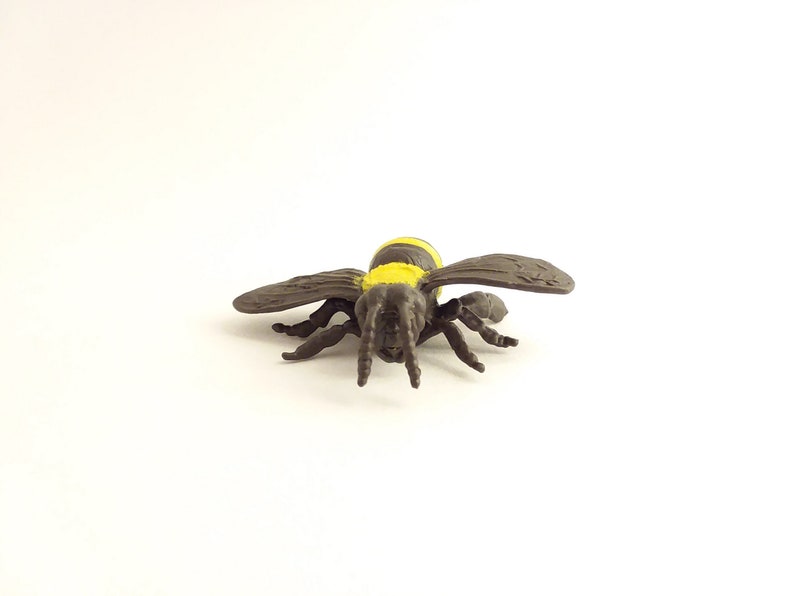 May include: A brown and yellow plastic toy bee with black eyes and six legs. The bee is facing the camera with its wings spread out.