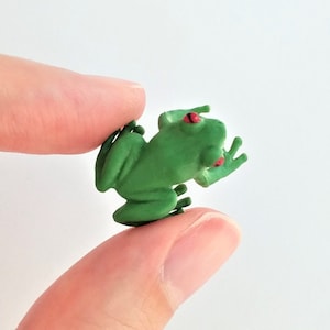 May include: A small green frog figurine with red eyes. The frog is posed with its legs bent and its head tilted up.