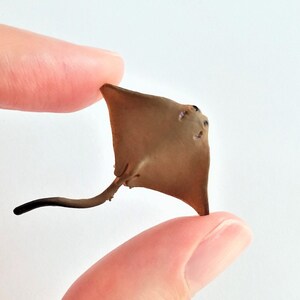May include: A small, brown stingray figurine with a long, thin tail. The figurine is held between two fingers.