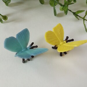 Peut inclure: Deux papillons en plastique, l'un bleu et l'autre jaune, avec des pattes et des antennes noires. Les papillons sont sur une surface blanche.