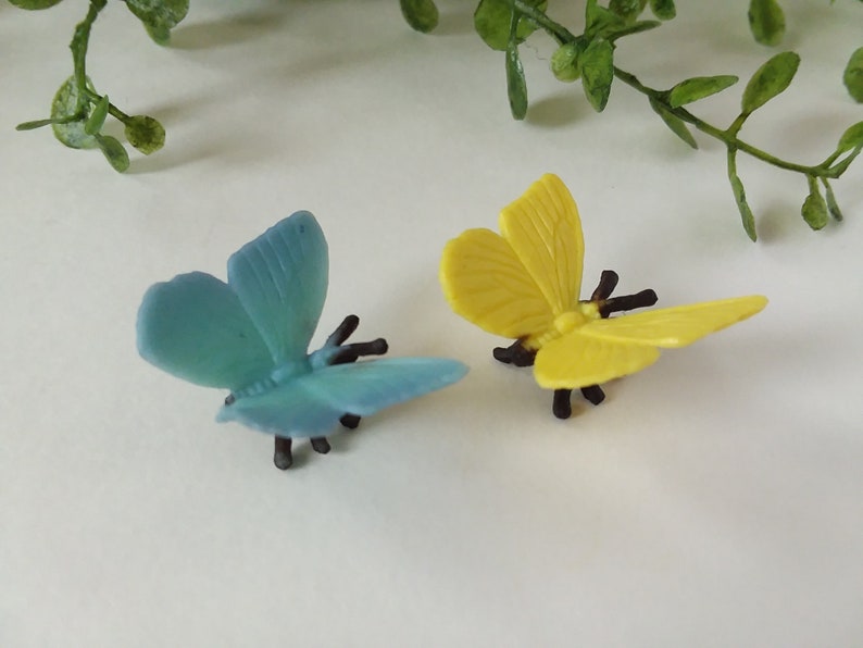 Peut inclure: Deux papillons en plastique, l'un bleu et l'autre jaune, avec des pattes et des antennes noires. Les papillons sont sur une surface blanche.