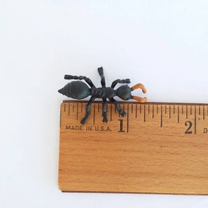 May include: A small, black plastic ant toy with orange pincers, positioned on a wooden ruler. The ant measures approximately 1 inch in length. The ruler is marked with inch increments and the text "MADE IN U.S."