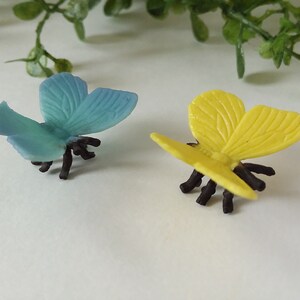 Peut inclure: Deux figurines de papillons en plastique, l'une bleue et l'autre jaune, avec des pattes noires. Les papillons sont pos&eacute;s avec leurs ailes ouvertes.