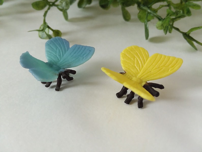 Peut inclure: Deux figurines de papillons en plastique, l'une bleue et l'autre jaune, avec des pattes noires. Les papillons sont pos&eacute;s avec leurs ailes ouvertes.