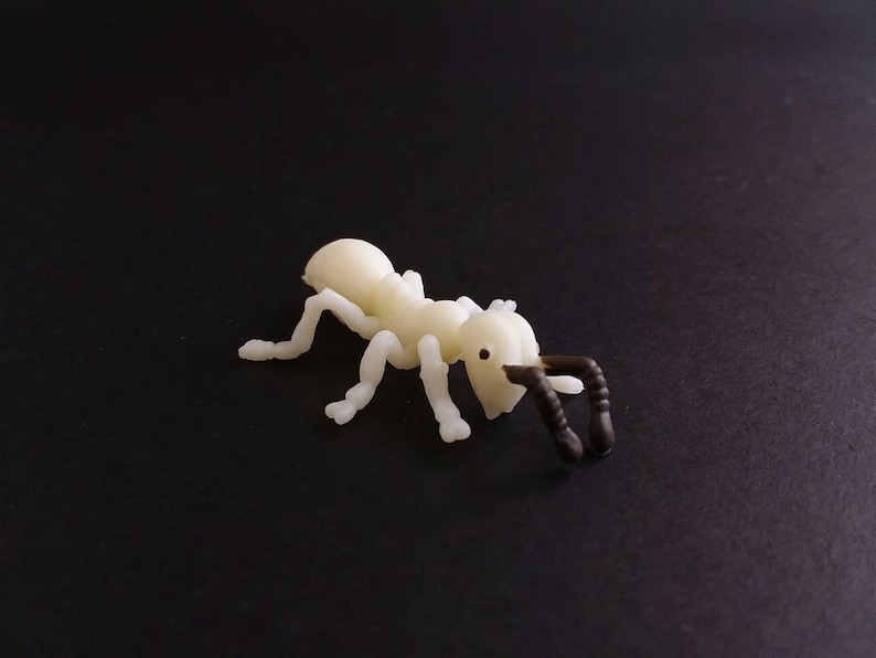 May include: A small, off-white plastic ant figurine with dark brown antennae and legs. The ant is positioned on a black surface, showcasing its detailed features and realistic design.