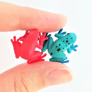 May include: Two small frog figurines, one red and one blue with black spots. The figurines are held in a person's hand.