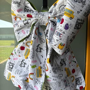 May include: A large, decorative bow with a white background and a repeating pattern of school-themed illustrations, including pencils, scissors, and school buses. The bow has a scalloped edge and green trim. Text includes "dream big!" and "you're the best!"