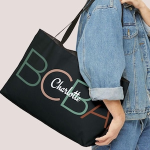 Puede incluir: Bolsa de tela negra con las letras "CBA" en verde, rosa y marrón. El nombre "Charlotte" está escrito en cursiva blanca sobre las letras.