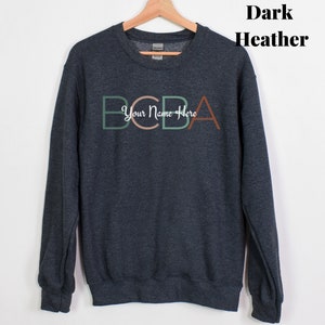 Peut inclure: Sweat-shirt gris chiné foncé avec le texte "BCBA Your Name Here" en vert, marron et rose.