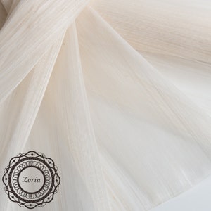 May include: Off-white fabric with a textured, flowing appearance. The fabric is draped and folded, creating soft shadows and highlights. A decorative logo with the word "Zoria" is in the lower left corner.