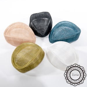 Op de afbeelding: Vijf gestructureerde fascinator-hoeden gerangschikt in een cirkelvormig patroon, met een geweven textuur in zwart, blauwgroen, beige, olijfgroen en wit. Elke hoed heeft een duidelijke druppel- of ovale vorm. Een cirkelvormig logo met de tekst Zoria is zichtbaar.