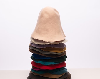 May include: A pile of felt hats in a range of colours, such as beige, purple, green, and red. The hats are stacked, with the beige hat at the top. The hats have a rounded, bell-like shape, and are made of felt.