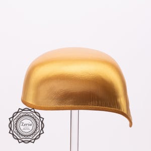 May include: A metallic gold satin head covering with a smooth, rounded dome shape and a structured, reinforced bottom edge. The item is displayed on a clear stand and features a circular black and white logo with the text Zoria in the corner.