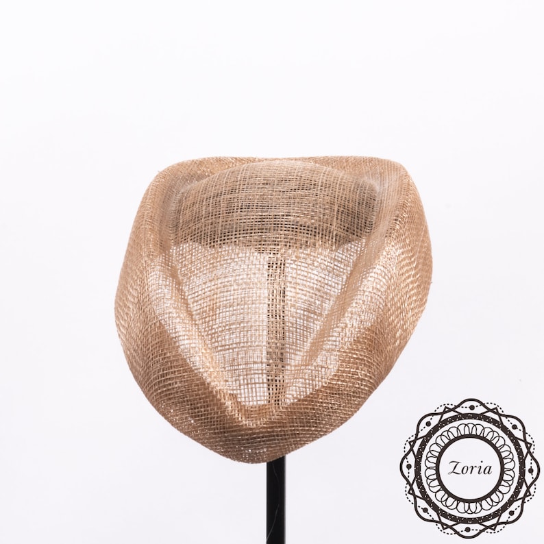 Op de afbeelding: Een gestructureerde, lichtbruine fascinatorhoed gemaakt van geweven, semi-transparant gaasmateriaal. Het hoofddeksel heeft een gebogen, sculpturaal silhouet met een prominente centrale koepel en een brede rand. Een logo met de tekst Zoria is zichtbaar.