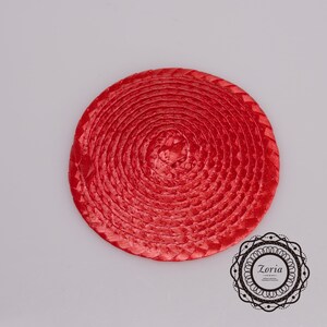 May include: A circular, bright red woven straw disc featuring a continuous spiral pattern. In the bottom right corner, there is a decorative black circular logo containing the text Zoria and smaller illegible text below it.