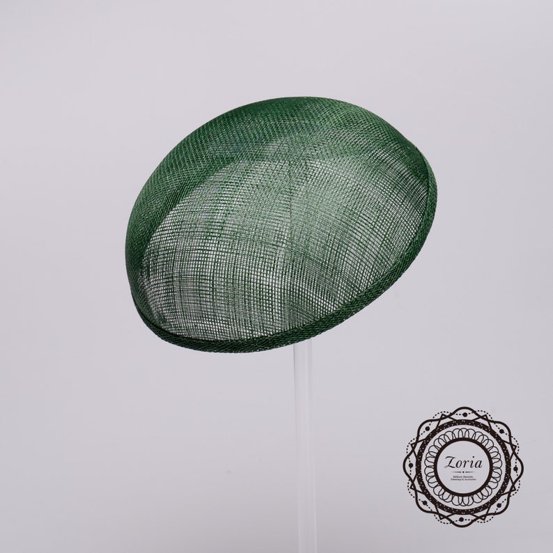 May include: A dark green, round, dome-shaped fascinator hat crafted from a stiff, woven mesh material. The hat features a smooth, curved silhouette and a structured brim edge. A circular logo with the text Zoria is visible in the corner.