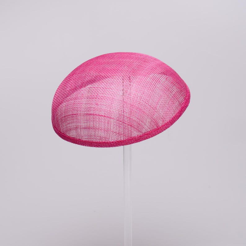 May include: A bright hot pink fascinator hat with a smooth, rounded dome shape. The hat is constructed from a fine, woven mesh material that features a visible cross-hatched texture throughout the surface, finished with a clean, bound edge.