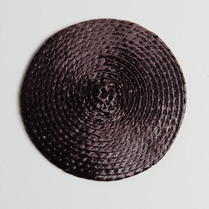 May include: A circular coaster featuring a dark brown, tightly woven, coiled fiber construction. The material is arranged in a continuous spiral pattern, radiating from the center to the outer edge, creating a textured, uniform surface.