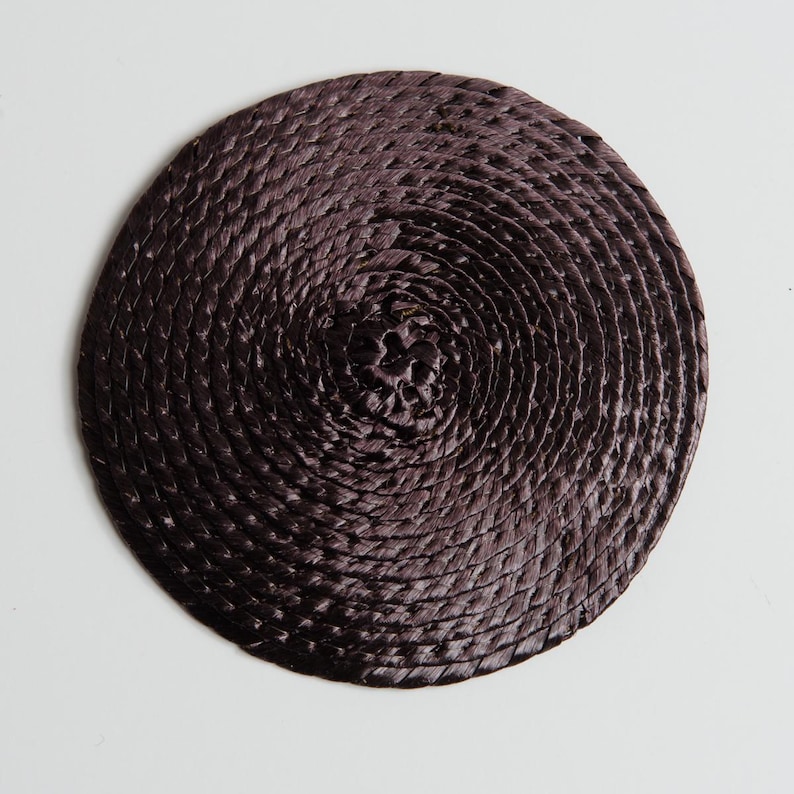 May include: A circular coaster featuring a dark brown, tightly woven, coiled fiber construction. The material is arranged in a continuous spiral pattern, radiating from the center to the outer edge, creating a textured, uniform surface.