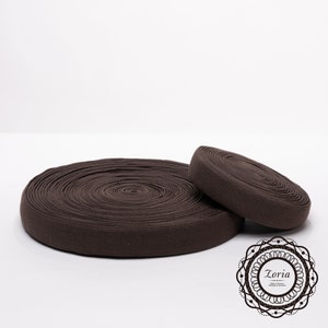 May include: A brown elastic band rolled up on a white surface. The elastic band is about 2 inches wide.