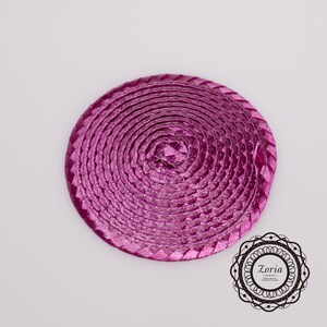 May include: A circular, magenta-colored woven straw disc featuring a continuous spiral pattern. The material has a shiny, metallic finish. In the bottom right corner, there is a decorative black circular logo containing the text Zoria.