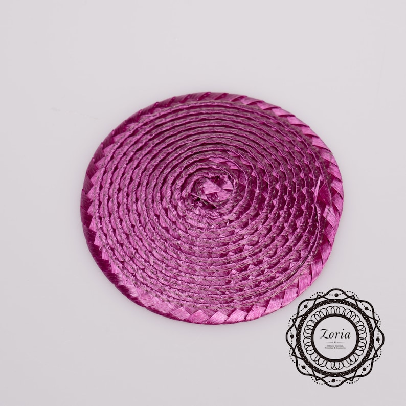 May include: A circular, magenta-colored woven straw disc featuring a continuous spiral pattern. The material has a shiny, metallic finish. In the bottom right corner, there is a decorative black circular logo containing the text Zoria.