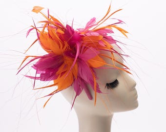 May include: A vibrant fascinator headpiece featuring a dense arrangement of bright orange and hot pink feathers. The feathers are layered and fanned out, creating a voluminous, textured look with long, thin wisps extending outwards.