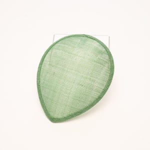 May include: A teardrop-shaped fascinator base in a light sage green color. The accessory features a fine, woven mesh texture throughout its surface, with a structured, reinforced border edge that maintains its distinct, pointed oval silhouette.