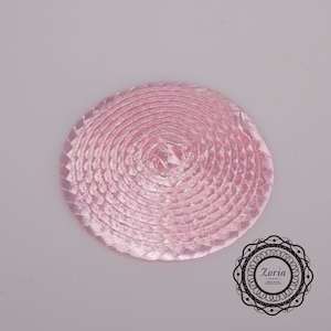 May include: A circular pink ribbon rosette featuring a tightly coiled, braided satin texture that spirals from the center outward. The design includes a decorative logo in the bottom right corner with the text Zoria.