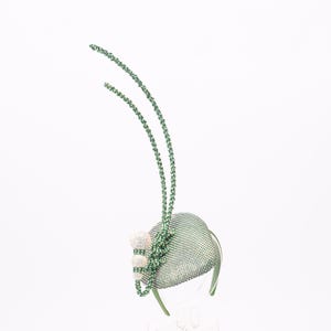 May include: A sage green fascinator headpiece featuring a structured, mesh-covered base. It is adorned with two long, slender, beaded antennae-like extensions and a cluster of three spherical, iridescent, light-reflecting beaded accents.