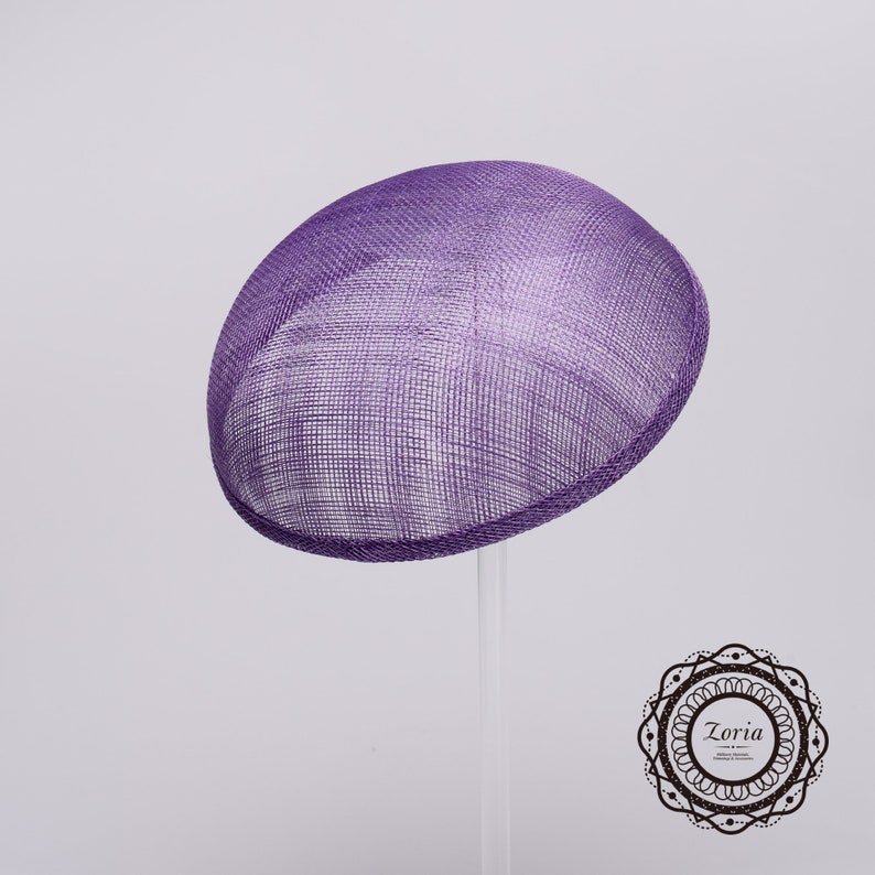 May include: A purple fascinator hat with a rounded, dome-shaped silhouette and a visible woven mesh texture. The hat features a clean, structured edge and is displayed on a clear stand. A circular logo with the text Zoria is visible.
