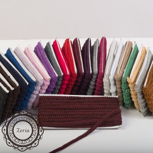May include: A collection of multi-colored crochet trim spools arranged in a semi-circle, featuring a prominent dark maroon spool in the foreground. Each spool displays a textured, braided pattern. The logo Zoria is visible in the bottom left corner.