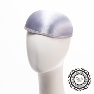 May include: A dome-shaped pillbox hat in a shimmering light lavender satin fabric. The hat features a structured, rounded crown and a thin, textured silver trim along the bottom edge. A circular logo with the text Zoria is visible in the corner.
