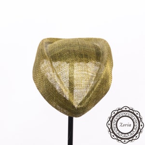 Op de afbeelding: Een goudkleurige fascinator-hoed met een gestructureerde, open geweven gaastextuur en een gebogen, sculpturaal silhouet. Het stuk is gemonteerd op een dunne zwarte standaard en heeft een rond logo in de rechterbenedenhoek met de tekst Zoria.